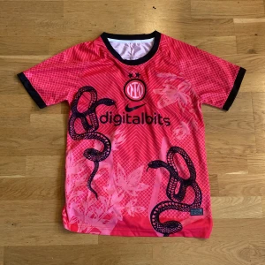 Inter Nike Dri-FIT rosa fotbollströja S - Inter fotbollströja från Nike i rosa med svarta detaljer och coolt ormmönster. Tröjan har korta ärmar, svart krage och är tillverkad i lätt Dri-FIT-material som andas. Digitalbits-logga på bröstet och klubbmärke med två stjärnor.