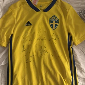 Svenska landslagets signerade fotbollströja från VM-laget 2018.  - Svenska landslaget matchtröja signerade av VM laget från 2018 seb Larsson, Olsen, Granqvist, mm. Gul fotbollströja från Adidas med blå detaljer och Sverige-emblem på bröstet. Tröjan har korta ärmar, V-ringad krage med blå kant och flera autografer på framsidan. Materialet är lätt och andas, perfekt för match eller samling. SVERIGE tryckt i nacken.