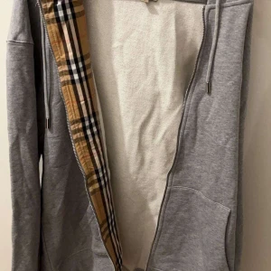 Grå hoodie med dragkedja - Grå hoodie  i bomull med klassiskt rutigt foder längs dragkedjan. Tröjan har huva med snörning, broderad logga på bröstet och två stora fickor framtill. 😊