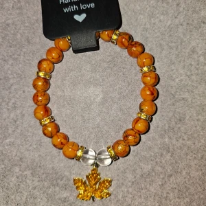 Orange pärlarmband med lövberlock - Unikt armband med orange marmorerade pärlor och guldfärgade detaljer. Armbandet har en snygg guldfärgad lönnlöv-berlock och två transparenta pärlor i mitten och 8st strassrondeller. Perfekt för dig som gillar höstiga färger och naturinspirerade smycken. Ca 17cm 