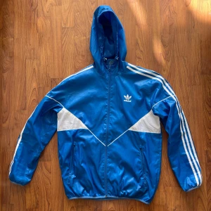 Blå Adidas vindjacka med huva M - Säljer en blå vindjacka från Adidas med klassiska vita ränder längs ärmarna och vita detaljer framtill. Jackan har huva med dragsko, hel dragkedja och två fickor med dragkedja. Materialet är lätt och syntetiskt, perfekt för blåsiga dagar.