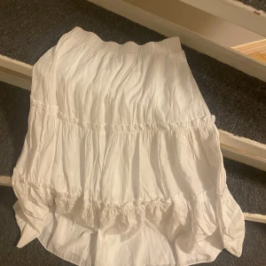 Vit volangkjol med resårmidja - Säljer en vit kortkjol med flera volanger och resår i midjan. Kjolen har en luftig och skön känsla, perfekt för varma dagar. Materialet är mjukt och lätt, och den har en feminin och avslappnad stil.