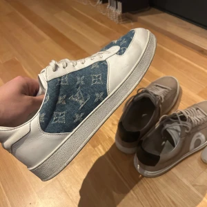 Louis Vuitton skor  - Säljer ett par Louis Vuitton sneakers med vit skinn och blå denim-paneler med klassiskt LV-monogram. Skorna har rund tå, snörning och platt sula. Kombinationen av skinn och denim ger en unik vibe och monogrammet gör dem riktigt lyxiga. Perfekt för dig som vill sticka ut med designer på fötterna. Skick 8,5/10 endast smutsiga 