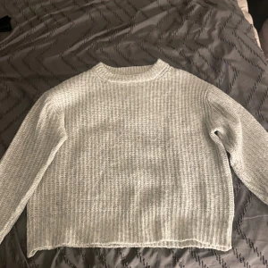 Ljusgrå stickad tröja med struktur - En ljusgrå stickad tröja med grov struktur och rund hals. Tröjan har långa ärmar och en avslappnad passform, perfekt för kyliga dagar. Enkel och stilren design som är lätt att matcha med olika outfits.