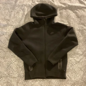 Svart hoodiejacka från Nike, storlek S Niketech fleece - Svart hoodiejacka från Nike med hel dragkedja framtill och diskret Nike-logga på bröstet. Jackan har huva, två fickor och är tillverkad i mjukt material som passar perfekt för chill eller träning.