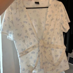 Vit blommig omlottblus med knyt - Säljer en söt vit omlottblus från Gina med ljusblått blommigt mönster. Blusen har korta ärmar, v-ringning och knyt band. Knappt använd och som ny! OBS köp helst inte via Bring!