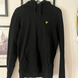 Svart hoodie från Lyle & Scott - Svart hoodie från Lyle & Scott med klassisk gul logga på bröstet. Tröjan har huva med snörning och en stor magficka framtill. Perfekt för dig som gillar en enkel och stilren look.