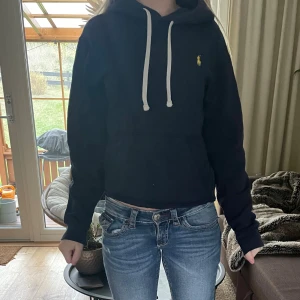 Mörk blå hoodie från Ralph Lauren - Mörk blå hoodie från Ralph Lauren med vit snörning och klassisk gul logga på bröstet. Tröjan har huva, känguruficka och långa ärmar. Perfekt för chill dagar och enkel att matcha med jeans eller joggers.