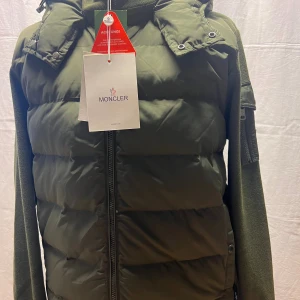 Olivgrön pufferjacka från Moncler - Snygg olivgrön pufferjacka från Moncler med stickade ärmar och huva. Jackan har dragkedja framtill, ficka med dragkedja på ärmen och Moncler-logga. Materialmix av polyester och stickat ger en modern vibe.
