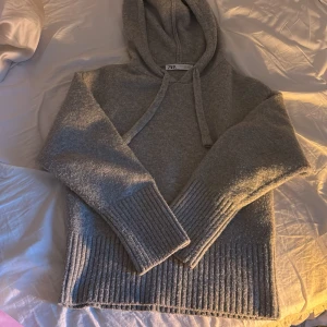 Grå stickad hoodie från Zara - Mysig grå hoodie från Zara i stickat material med huva och snörning. Tröjan har långa ärmar och breda ribbade muddar vid ärmslut och nederkant. Perfekt för kyliga dagar och enkel att styla med jeans eller mjukisbyxor.