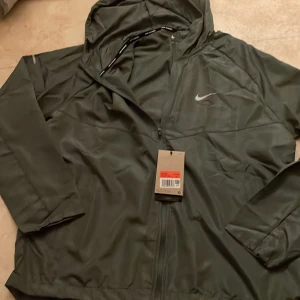Grå nike Miller - Nike vindjacka i mörkgrönt med huva och dragkedja framtill. Jackan har diskret Nike-logga på bröstet, innerficka med dragkedja och meshfoder. Perfekt för löpning eller utomhusaktiviteter. Snygg och sportig design med reflekterande detaljer på ärmarna.