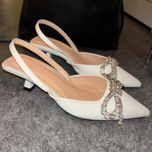 Två par vita klackskor med strass - Säljer två par vita klackskor. Ena paret har spetsig tå, låg klack och glittrig strassrosett framtill, med öppen häl och rem. Andra paret är pumps med hög klack, spetsig tå och täckt av små pärlor och strass för extra bling. Perfekta för dig som vill sticka ut.