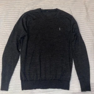 Ralph lauren merinoull tröja - Mörkgrå stickad tröja från Polo Ralph Lauren med klassisk rund halsringning och broderad logga på bröstet. Tröjan är gjord i 100% merinoull. Storleken är M men passar S