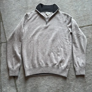 Grå half zip tröja från Hartford - Snygg grå långärmad tröja från Hartford med ribbad svart insida på kragen och half zip-dragkedja. Perfekt för dig som gillar en clean och chill stil. Mjuk och skön i kashmir, passar till jeans eller chinos.