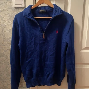 Blå stickad tröja Polo Ralph Lauren - Mörkblå stickad tröja från Polo Ralph Lauren med halv dragkedja och ribbad krage. Klassisk röd logga broderad på bröstet. Långärmad modell med ribbade muddar och nederkant. Perfekt för lager-på-lager under kyliga dagar.