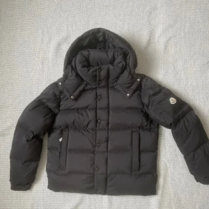 Moncler Vezere - Svart dunjacka från Moncler med avtagbar huva, klassisk Moncler-logga på ärmen och quiltad design. Jackan har dragkedja och knappar framtill, samt praktiska fickor. Insidan har ett serietidningsmönster och Moncler-etikett. Perfekt för kalla dagar.
