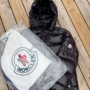 Moncler Maya - Säljer en svart pufferjacka från Moncler med glansig finish, huva och dragkedja framtill. Jackan har Moncler-logga på ärmen och praktisk ficka med lock. Insidan har ett unikt serietryck. Perfekt för kalla dagar och riktigt snygg streetstil. Kvitto och Dustbag finns. Size 2 = M. Jackan är använd sparsamt och försiktigt.