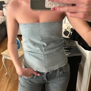 Ljusblå jeans tubtopp med dragkedja - Säljer en snygg ljusblå tubtopp i jeansmaterial med rå kant nertill och vit dragkedja på sidan. Toppen är axelbandslös och har en tight passform som ger en cool och trendig look. Perfekt att matcha med jeans för en denim-on-denim vibe.