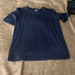 Blå t-shirt med tryck, storlek S - Snygg marinblå t-shirt i mjuk och stretchig polyester med coolt vitt tryck på ryggen: 'Forever Chasing Sunsets' med palm och blomma. Perfekt för dig som gillar chill vibes och grafiska prints. Rund hals och korta ärmar, passar dig som vill ha en relaxed stil.