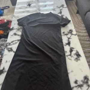 Svart lång t-shirtklänning - En enkel svart långklänning med t-shirtmodell och korta ärmar. Klänningen har rak passform och är tillverkad i mjukt bomullstyg, perfekt för en chill och avslappnad look. Stilren och lätt att matcha med sneakers eller boots.