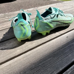 Nike Mercurial fotbollsskor turkos - Nike Mercurial fotbollsskor i en fräsch turkos och limegrön färg med svarta detaljer. Skorna har snörning, platt sula med dobbar och är tillverkade i syntetmaterial för bästa grepp på planen. Perfekta för dig som vill sticka ut på fotbollsplanen.