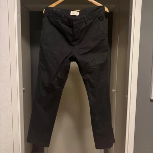 Svarta chinos från GANT - Snygga svarta chinos från GANT med klassisk rak passform och knappstängning fram. Byxorna har diskreta fickor och är perfekta för en stilren look. Materialet känns mjukt och bekvämt mot huden. Passar dig som gillar enkel och tidlös stil.