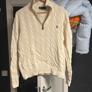 Vit kabelstickad tröja Polo Ralph Lauren - Snygg kabelstickad tröja från Polo Ralph Lauren i vit färg med half zip och broderad logga på bröstet. Tröjan har långärmad passform, ribbade muddar och är gjord i mjuk bomull. Perfekt för dig som gillar klassisk stil med en modern touch.