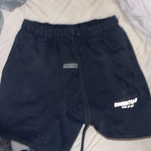 Svarta shorts från Essentials Fear of God - Svarta shorts från Essentials Fear of God med vit logga på vänster ben och snygg snörning i midjan. Mjukt material och relaxed fit, perfekt för chill dagar. Elastisk midja och bakficka för extra stilpoäng.