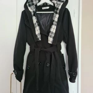 Svart kappa med dubbelknäppning  - Svart kappa med dubbelknäppning och bälte i midjan. Huva med svartvitt rutigt foder som ger en cool kontrast. Kappan har långa ärmar med knappdetaljer och är fodrad för extra värme. Perfekt för dig som gillar stilrena och klassiska plagg med en twist.
