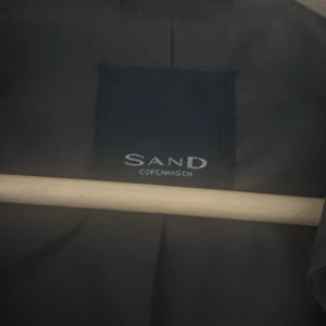 Svart trenchcoat från Sand Copenhagen - Snygg mörkgrön / militärgrön trenchcoat från Sand Copenhagen med klassisk krage, långa ärmar och cool asymmetrisk dragkedja framtill. Jackan har stilrena detaljer vid ärmsluten och är perfekt för dig som gillar minimalistisk design. Materialet känns slätt och lätt.