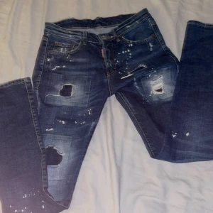 Dsquared2 blå distressed jeans - Snygga blå jeans från Dsquared2 med coola slitningar, hål och färgstänk för en riktigt edgy look. Klassisk femficksmodell med gylf och knapp, samt Dsquared2-logga vid fickan. Jeansen har en rak passform och är tillverkade i jeansmaterial.