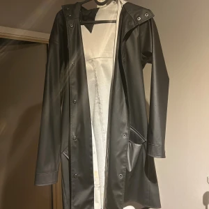 Svart regnjacka med huva och snörning - Säljer en svart regnjacka med huva och snören. Jackan är lång (på mig till strax över knäna jag är 169cm)  har en lite slits i bak, tryckknappar framtill och två stora fickor. Materialet är slätt och vattenavvisande. Bikbok märket är en triangel ibak som syns så de kliar inte eller något sånt