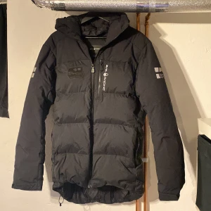 Svart pufferjacka från Sail Racing - Svart pufferjacka från Sail Racing med huva och dragkedja framtill. Jackan har logga på bröstet och ärmen, samt diskreta reflexdetaljer. Fylld för extra värme och perfekt för kalla dagar. Snygg, sportig look med normal passform.