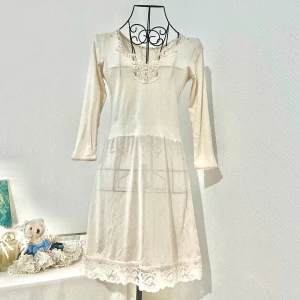 Långarmad nattlinne i ljusbeige/ljusgul - Långarmad nattlinne i ljusbeige/ljusgul med fina spetsdetaljer. Den nedre delen är något genomskinlig och har liten mörkare färg (typ ljusorange) men självklart ingen fläck. Passar bra storlek 34-36. Dockan har storlek 34.