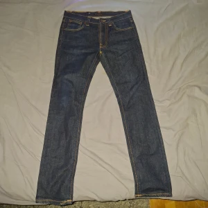 Nudie Jeans blå jeans 30/30 - Snygga blå jeans från Nudie Jeans i modellen 30/30. Jeansen är tillverkade av 100% ekologisk bomull och har klassiska orangea sömmar samt Nudie-logga på bakfickorna. Raka ben och normal passform, perfekt för dig som gillar stilrena och tidlösa jeans. Inga defekter, säljs pga för små för mig, helt nya