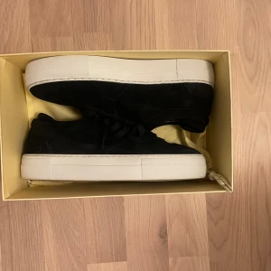 Svarta sneakers från Axel Arigato - Snygga svarta sneakers från Axel Arigato i mocka med vit platt sula. Skorna har snörning och diskret guldfärgad logga på sidan. Perfekta för dig som gillar stilrena och trendiga skor med en lyxig känsla.