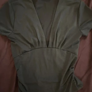 Grön v ringad topp  - Snygg grön topp med djup v-ringning och korta ärmar. Tröjan  har en markerad midja och lätt rynkade detaljer vid bysten för en cool och modern look. Perfekt för dig som gillar stilrena och enkla plagg.