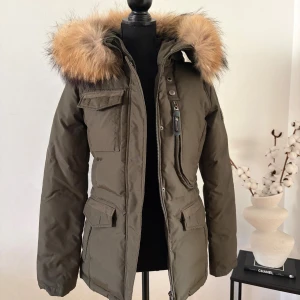 Olivgrön Hollies Livigno äkta päls - Snygg olivgrön jacka från Hollies med stor fluffig pälskant på huvan. Jackan har flera fickor med lock, dragkedja och knappar framtill samt patch på ärmen. Perfekt för kalla dagar med sin varma och stilrena look. Jackan är i nyskick! 