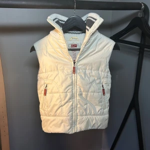 Vit quiltad dunväst från Carry - Snygg vit dunväst från Carry med quiltad framsida i glansig polyester och dragkedjor framtill. Baksidan och kragen är stickad i grått och vitt randigt mönster, vilket ger en unik look. Perfekt för lager-på-lager och extra värme under kyliga dagar. 