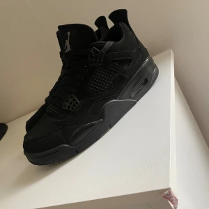 Svarta Nike Air Jordan 4 sneakers - Säljer ett par helsvarta Nike Air Jordan 4 sneakers med klassisk Jumpman-logga på plösen och hälen. Skorna har snörning, meshpaneler och detaljer i mocka och syntet. Snygg och sportig siluett som passar perfekt till streetwear.
