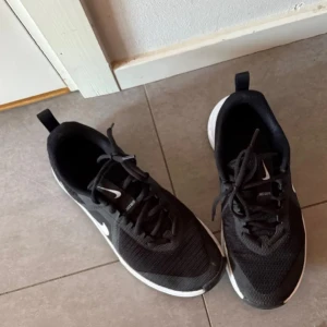 Svarta Nike sneakers med vit sula - Svarta Nike sneakers med vit sula och klassisk swoosh på sidan. Skorna har snörning och är tillverkade i mesh och syntetmaterial för bra ventilation. Rund tå och platt sula gör dem perfekta för en aktiv livsstil. Snygg och sportig design som passar till det mesta.