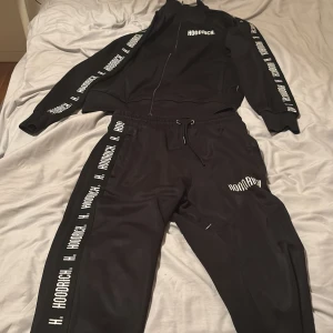 Svarta Hoodrich mjukisbyxor med tryck - Svarta mjukisbyxor från Hoodrich med vit logotyp längs benen och på höften. Byxorna har snörning i midjan och en relaxed passform. Materialet är mjukt och skönt, perfekt för chill eller träning. Snygg streetstil med tydliga tryck längs sidorna. 300kr per plagg.