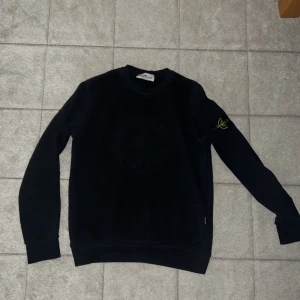 Svart Stone Island sweatshirt M - Svart sweatshirt från Stone Island i storlek M. Klassisk rund hals och lång ärm med ikonisk logotyp-patch på vänster arm. Tröjan har ett diskret broderat emblem på bröstet och är gjord i mjuk bomull för skön komfort. Perfekt för dig som gillar streetwear och snygga detaljer.passar stl S också