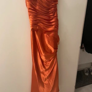 Lång orange balklänning med blomdetalj - Snygg långklänning i glansigt orange satinliknande material med draperad design och en stor blomma på axeln. Klänningen har smala axelband, figurnära passform och dragkedja i sidan. Perfekt för bal eller speciella tillfällen.