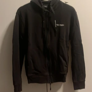 Svart hoodie från Armani Exchange - Svart hoodie från Armani Exchange med dragkedja hela vägen och snörning i huvan. Tröjan har diskret logga på bröstet och tvåvägsdragkedja. Klassisk passform och mjukt material som känns skönt mot huden.