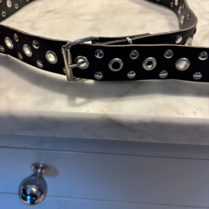 Svart bälte med silvernitar, 75 cm - Svart bälte med dubbla rader av stora och små silverfärgade nitar och hål längs hela längden. Snyggt metallspänne i silver. Perfekt för att ge din outfit en edgy vibe. Bältet är ca 75 cm långt och har en klassisk rak form. Aldrig använt