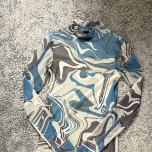 Blå och grå mönstrad långärmad klänning - Säljer en långärmad klänning med coolt abstrakt mönster i blått, grått och vitt. Klänningen har hög hals och är figurnära med ett stretchigt material som känns bekvämt. Perfekt för dig som gillar unika prints och vill sticka ut.