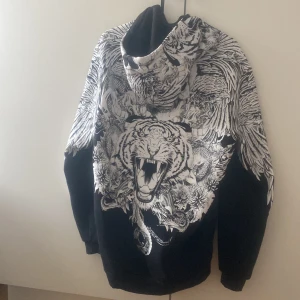 Svart hoodie med tigertryck - Svart hoodie med grymt detaljerat vitt tryck av en rytande tiger och coola fågelmotiv på ryggen och ärmarna. Har huva med snörning och half zip framtill. Materialet känns som mjuk bomull, perfekt för dig som gillar statement-plagg.