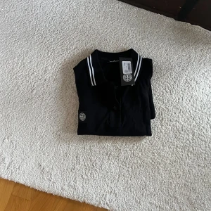 Svart Stone Island pikétröja - Svart pikétröja från Stone Island med klassisk krage och vita ränder på krage och ärmslut. Tröjan har korta ärmar, knappslå framtill och ikonisk logotyp på bröstet. Tillverkad i 100% bomull för en skön känsla.