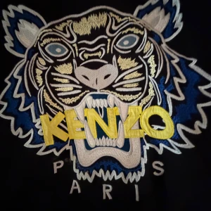 Svart Kenzo sweatshirt med tiger XS - Svart sweatshirt från Kenzo i storlek XS med ikonisk broderad tiger på bröstet i blått, gult, vitt och grått. Tröjan har rund hals, ribbade muddar och lång ärm. Perfekt statement-plagg med coolt Paris-tryck under tigern.